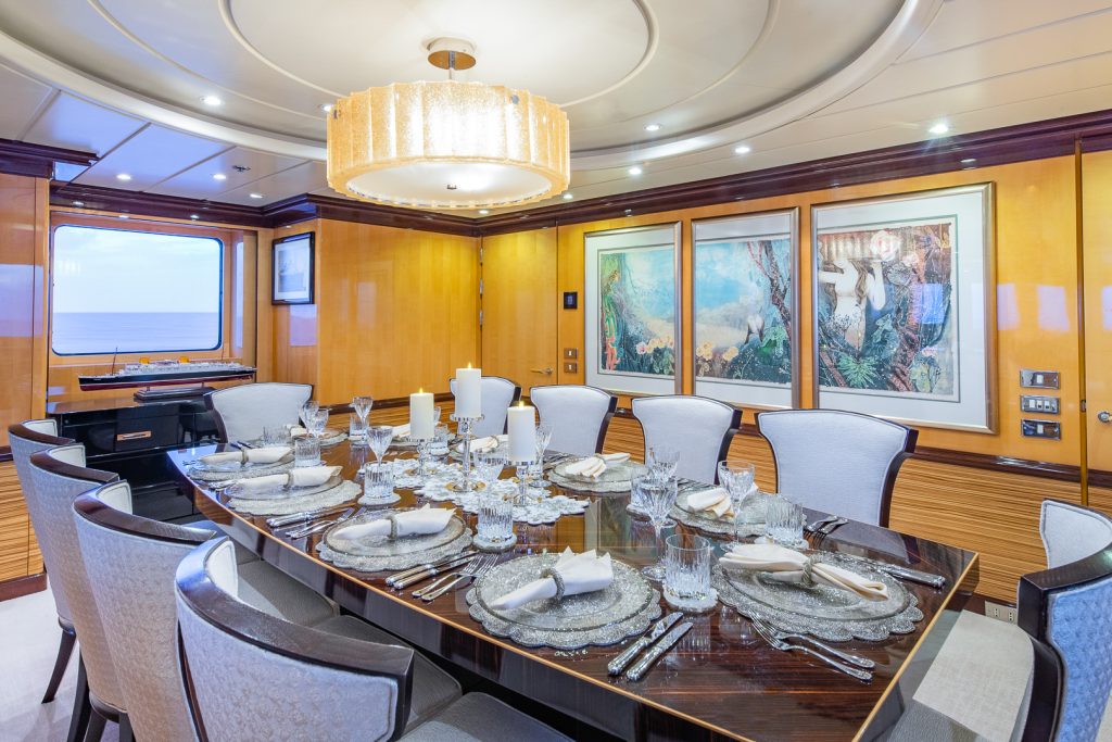 Elegant yacht dining room with a glossy wooden table set for eight, featuring crystal glassware, white plates, and napkins. The room has soft lighting, light wood walls, and framed art, with ocean visible through a window.