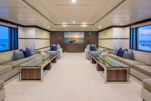 Luxurious yacht interior with beige sofas, blue cushions, glass coffee tables, and large windows showing the ocean. A painting of a boat at sunset hangs on the far wall, and subtle lighting creates an elegant atmosphere.