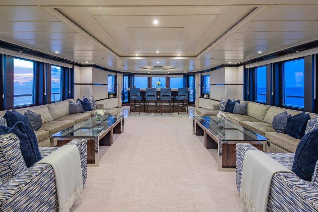 Spacious yacht interior with large sofas, blue and beige decor, glass coffee tables, and wide windows showing ocean views. A dining table with chairs is at the far end, under soft ceiling lights.
