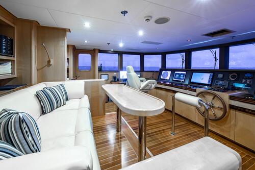 Spacious yacht bridge with modern navigation panels, large windows, a central captain’s chair, wooden flooring, a white leather sofa with striped pillows, bookshelves, and natural light from outside.