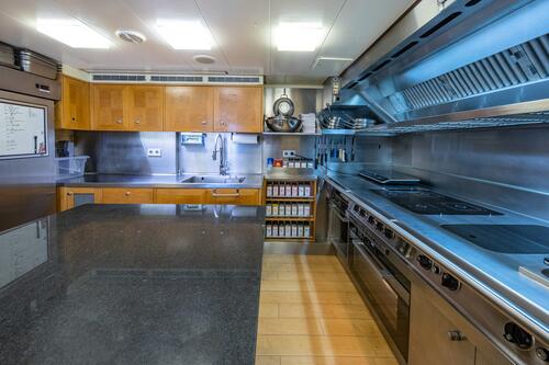 A modern commercial kitchen with stainless steel appliances, induction stoves, wooden cabinets, spice jars, a large sink, and a granite countertop. The space is clean, organized, and well-lit.