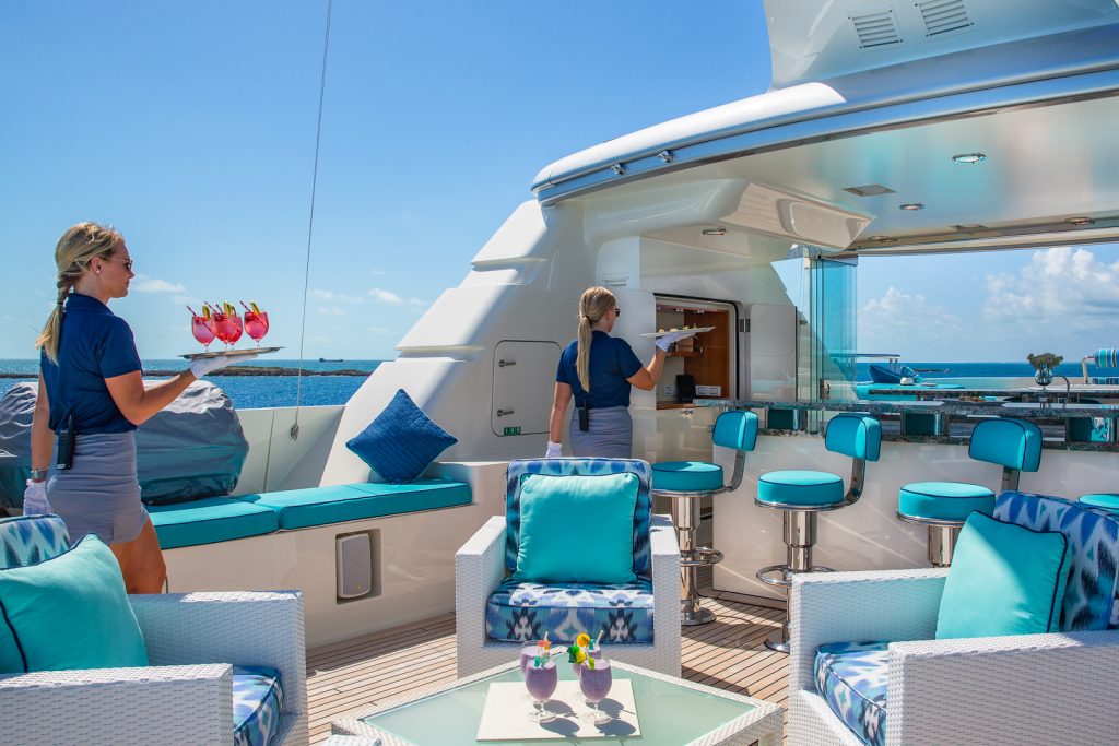 A woman in a blue shirt and gray skirt carries drinks on a tray toward an outdoor yacht bar, where another woman prepares glasses. The yacht features turquoise seating and ocean views under a sunny blue sky.