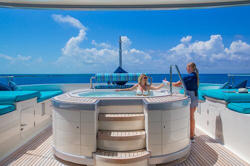 A woman relaxes in a yacht hot tub while another woman hands her a drink; the deck has blue seating, with the ocean and a bright blue sky in the background.
