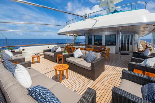 Spacious yacht deck with wicker sofas, tables, and cushioned chairs, set under a partly sunny sky and overlooking the ocean. Dining table and spiral staircase are visible near the cabin entrance.
