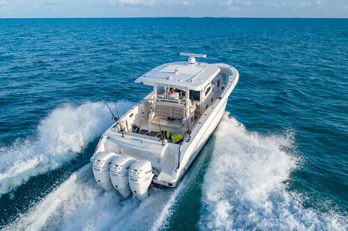 A white motorboat with three engines speeds across clear blue ocean water, leaving a trail of foamy waves behind it under a partly cloudy sky.