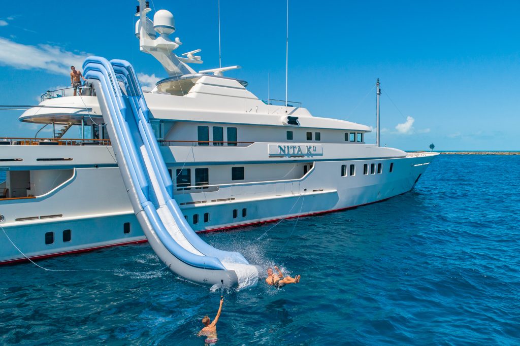 A large white yacht with a blue inflatable slide is anchored in the ocean. Three people are sliding down and splashing into the water on a sunny, clear day.