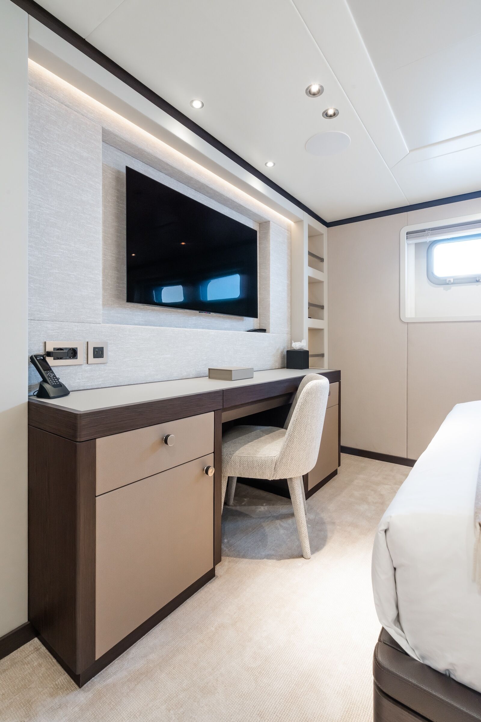 Modern bedroom workspace with a built-in desk, beige chair, wall-mounted TV, shelves, and a small window letting in natural light. The room has a clean, minimalist design with neutral colors.