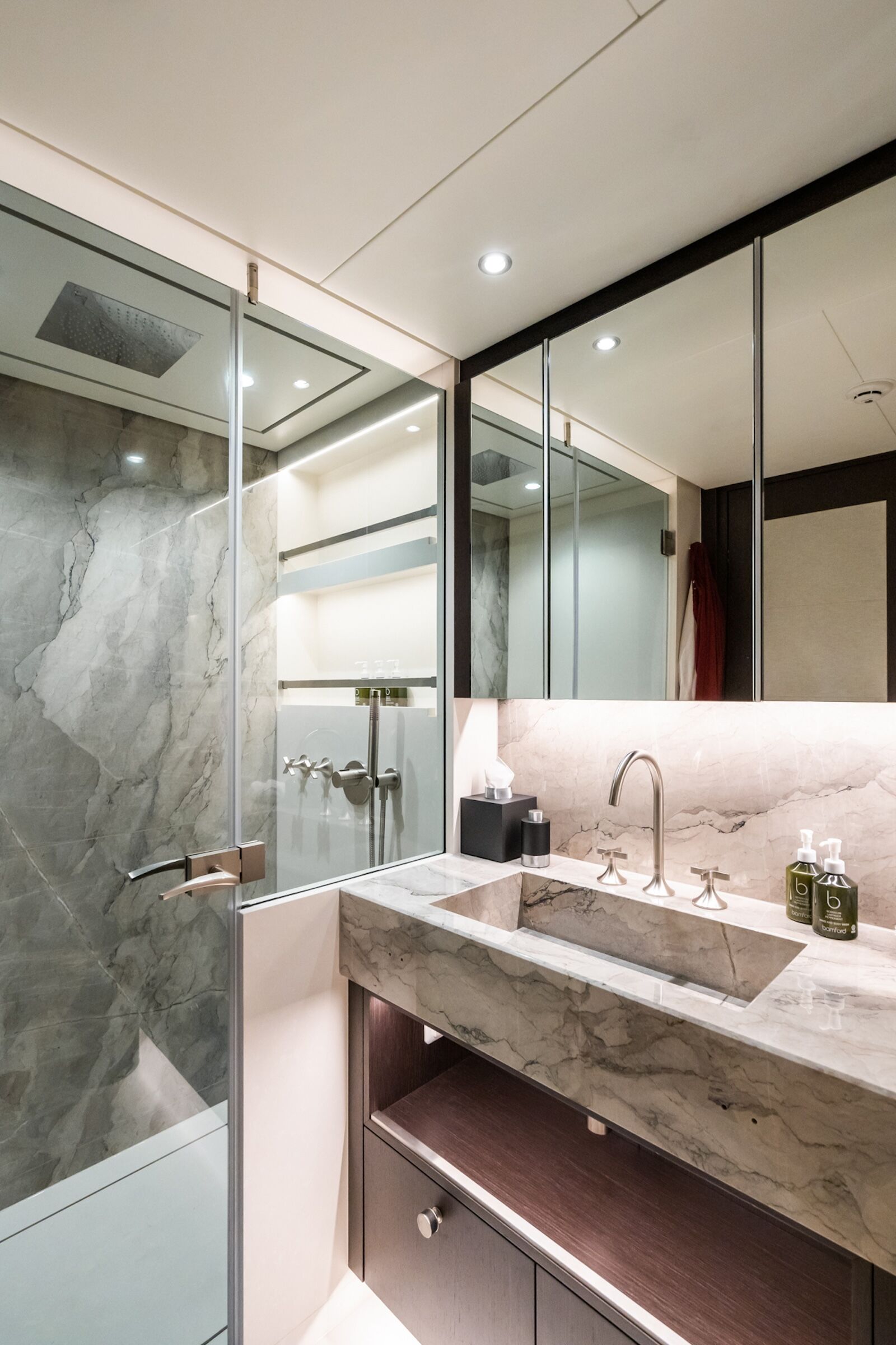A modern bathroom with a glass-enclosed shower, marble walls, a marble sink countertop, built-in shelves, a large mirror, and under-sink storage. The space is well-lit with recessed ceiling lights.