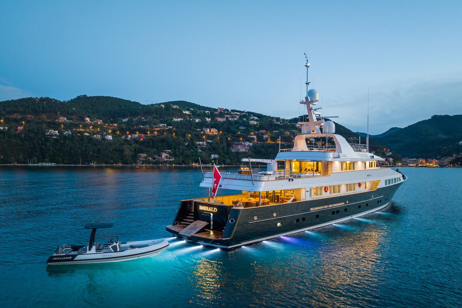 A large luxury yacht with glowing underwater lights is anchored near a coastal town at dusk, with a smaller motorboat tied beside it and hills dotted with houses in the background.