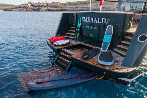 A yacht named Emerald Valletta has several water sports boards, a red life jacket, and other gear arranged on its rear swim platform, moored on calm blue water with a rocky coast in the background.