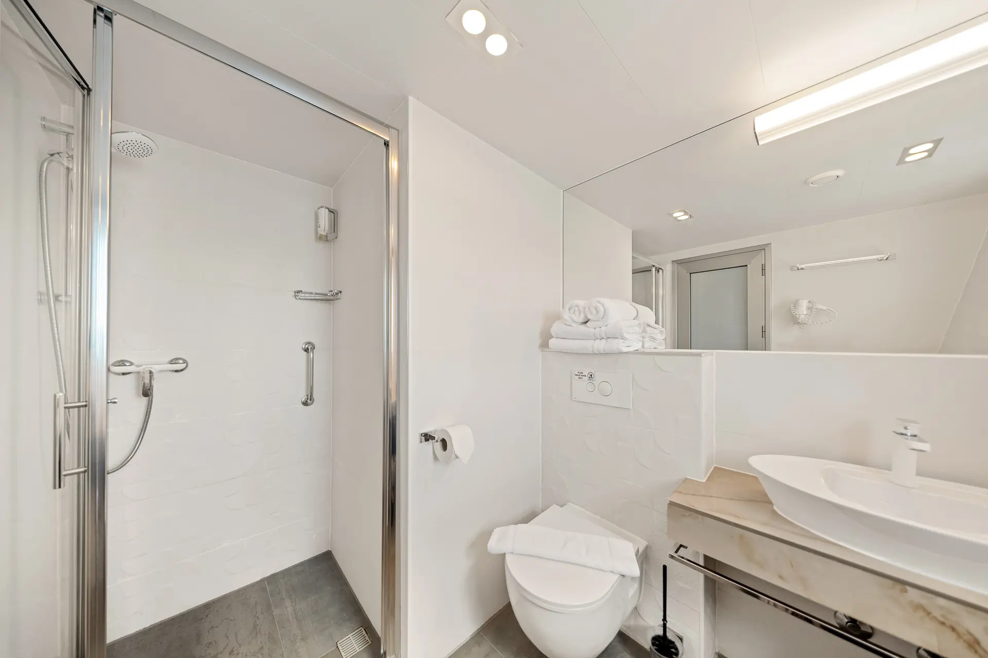 Modern, clean bathroom with a glass-enclosed shower, white walls, wall-mounted toilet with folded towels on top, a vessel sink on a marble counter, and a large mirror reflecting the space.