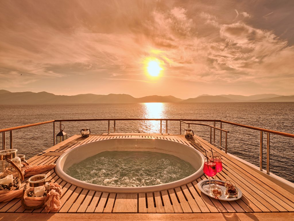 A round hot tub on a yacht deck overlooks a calm sea at sunset, with mountains in the distance. Drinks and snacks sit on a wooden table beside the tub, creating a relaxing and luxurious atmosphere.