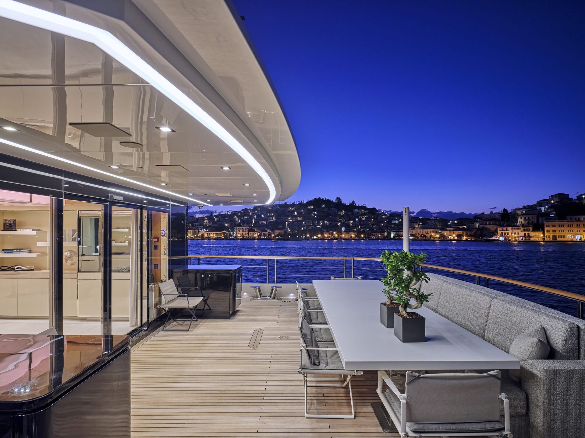 Modern yacht deck at dusk with a long dining table, cushioned seating, and potted plants, overlooking calm water and a cityscape with illuminated buildings and hills in the background.