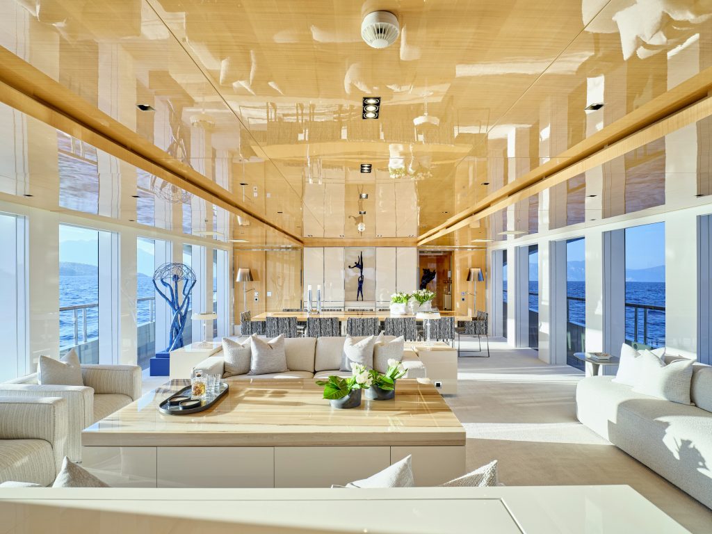 Spacious, modern yacht interior with glossy wooden ceiling, large windows offering ocean views, white sofas, green plants, and contemporary sculptures. Dining area and seating are arranged in an open, bright layout.