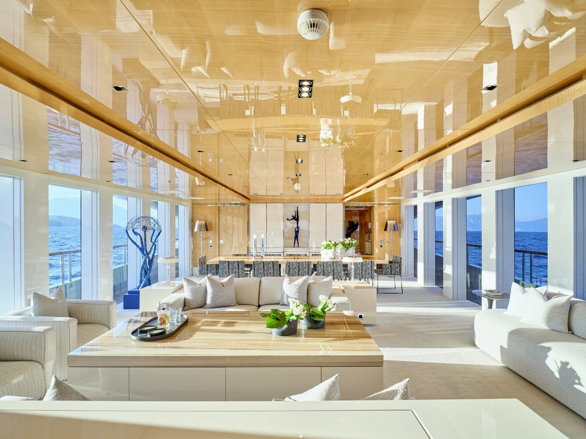 Spacious, modern yacht interior with glossy wooden ceiling, large windows offering ocean views, white sofas, green plants, and contemporary sculptures. Dining area and seating are arranged in an open, bright layout.