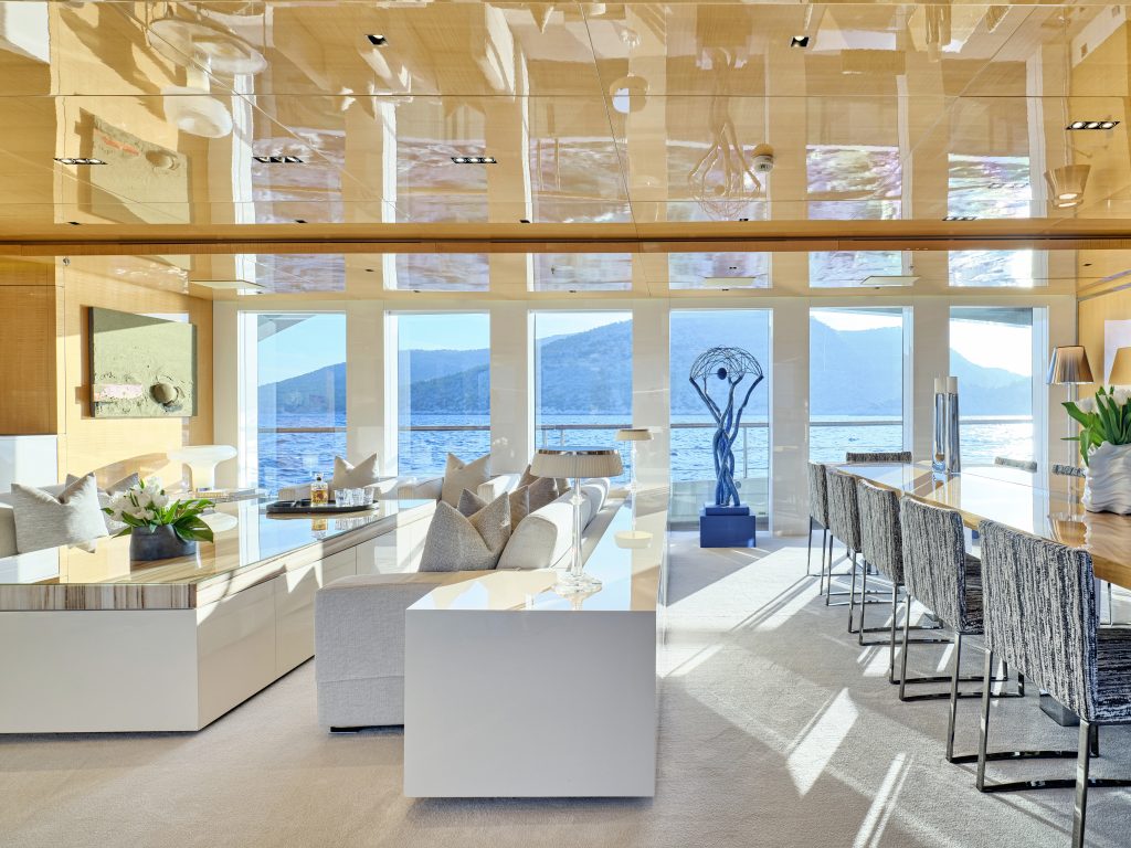 Modern, sunlit living and dining area with neutral tones, large windows overlooking water and mountains, a reflective ceiling, contemporary furniture, and a blue sculpture on the balcony outside.