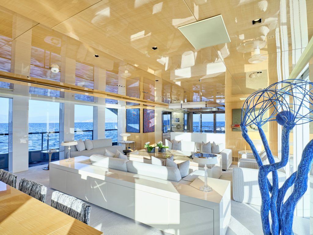Modern, sunlit living room with light wood walls and ceiling, large windows overlooking the ocean, white sofas, glass coffee table, and a blue abstract sculpture in the foreground.