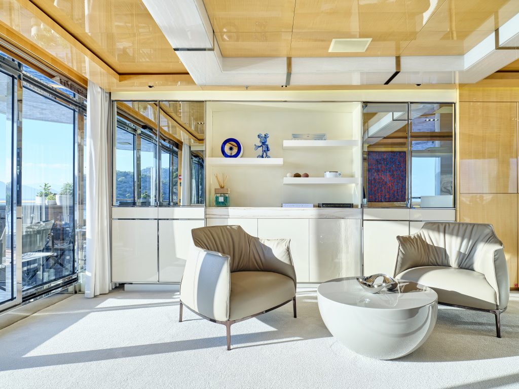 Modern living room with large windows, two gray armchairs, a round white coffee table, built-in shelves with decorative items, and natural light filling the bright, minimalist space.