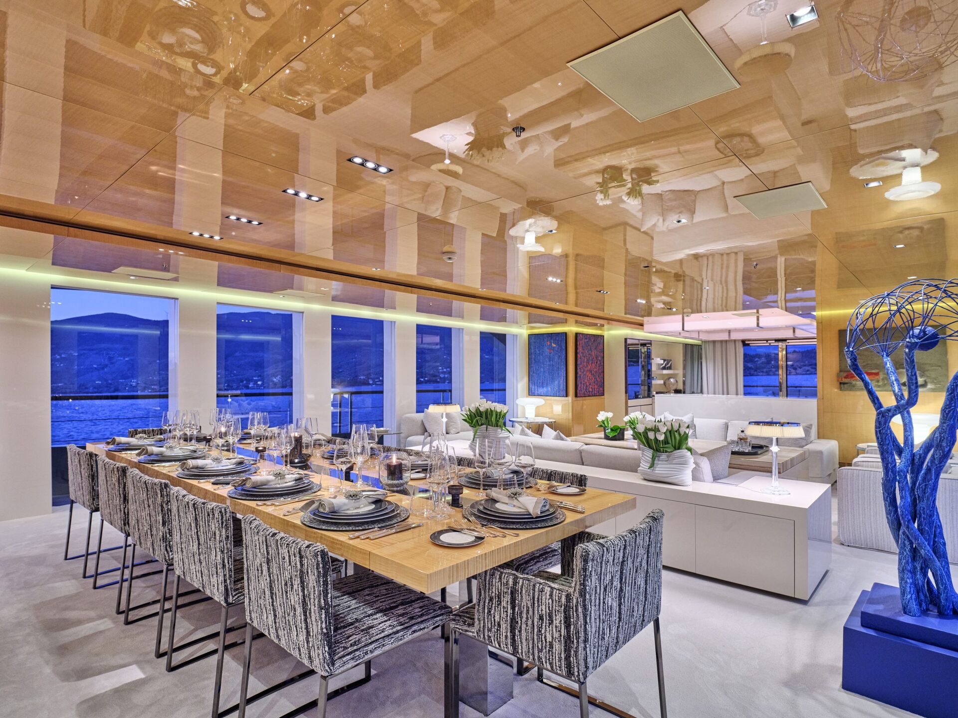 Modern dining and living area with a long table set for a meal, stylish gray chairs, large windows showing a waterfront view at dusk, and contemporary décor including a blue sculpture and reflective ceiling.