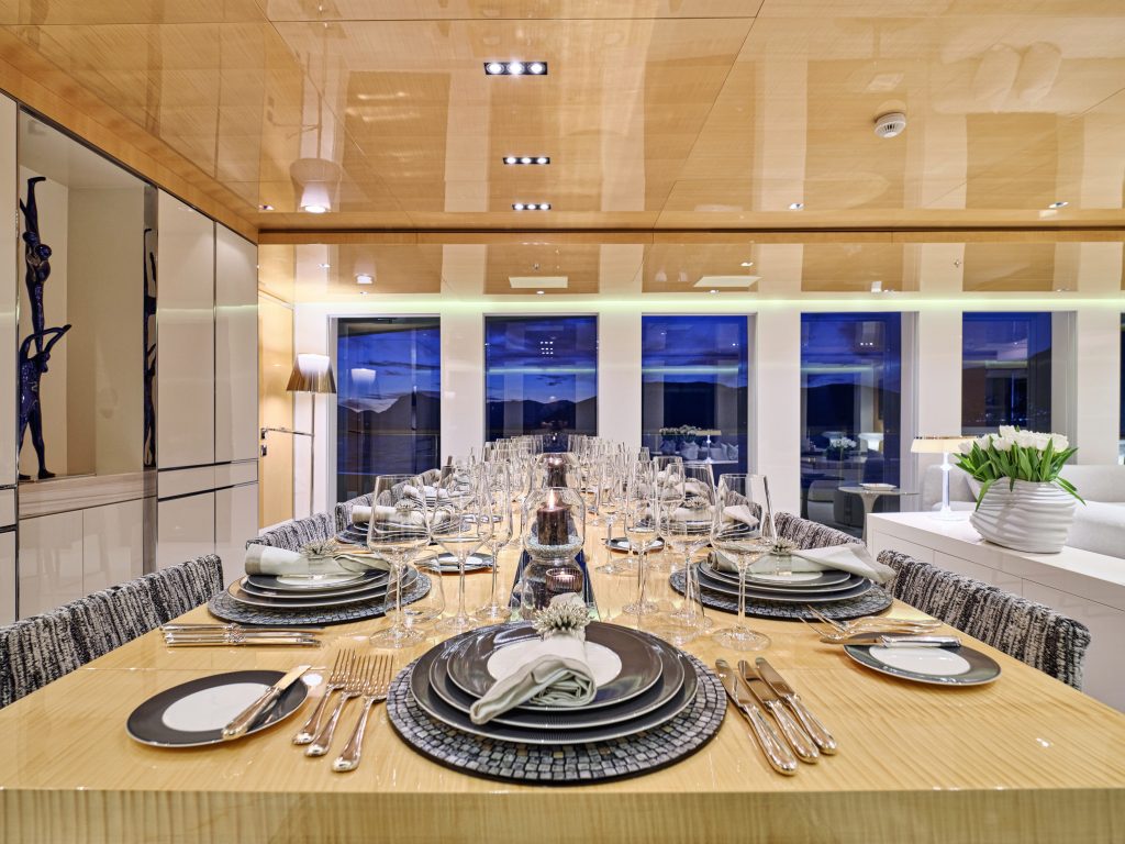 A modern, elegant dining room with a long wooden table set for a formal dinner, featuring neatly arranged plates, napkins, glasses, and silverware. Large windows show an evening view outside.