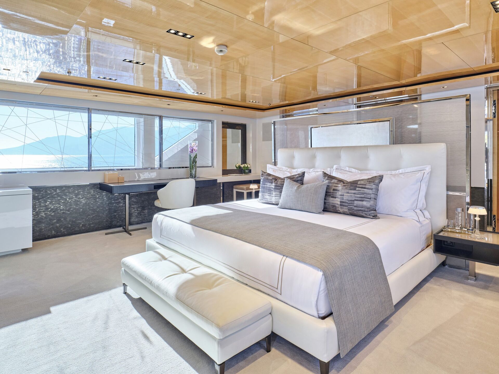 Luxurious modern bedroom with a large bed, padded bench, desk, and floor-to-ceiling windows offering a scenic water view. The room features light wood accents and contemporary decor.