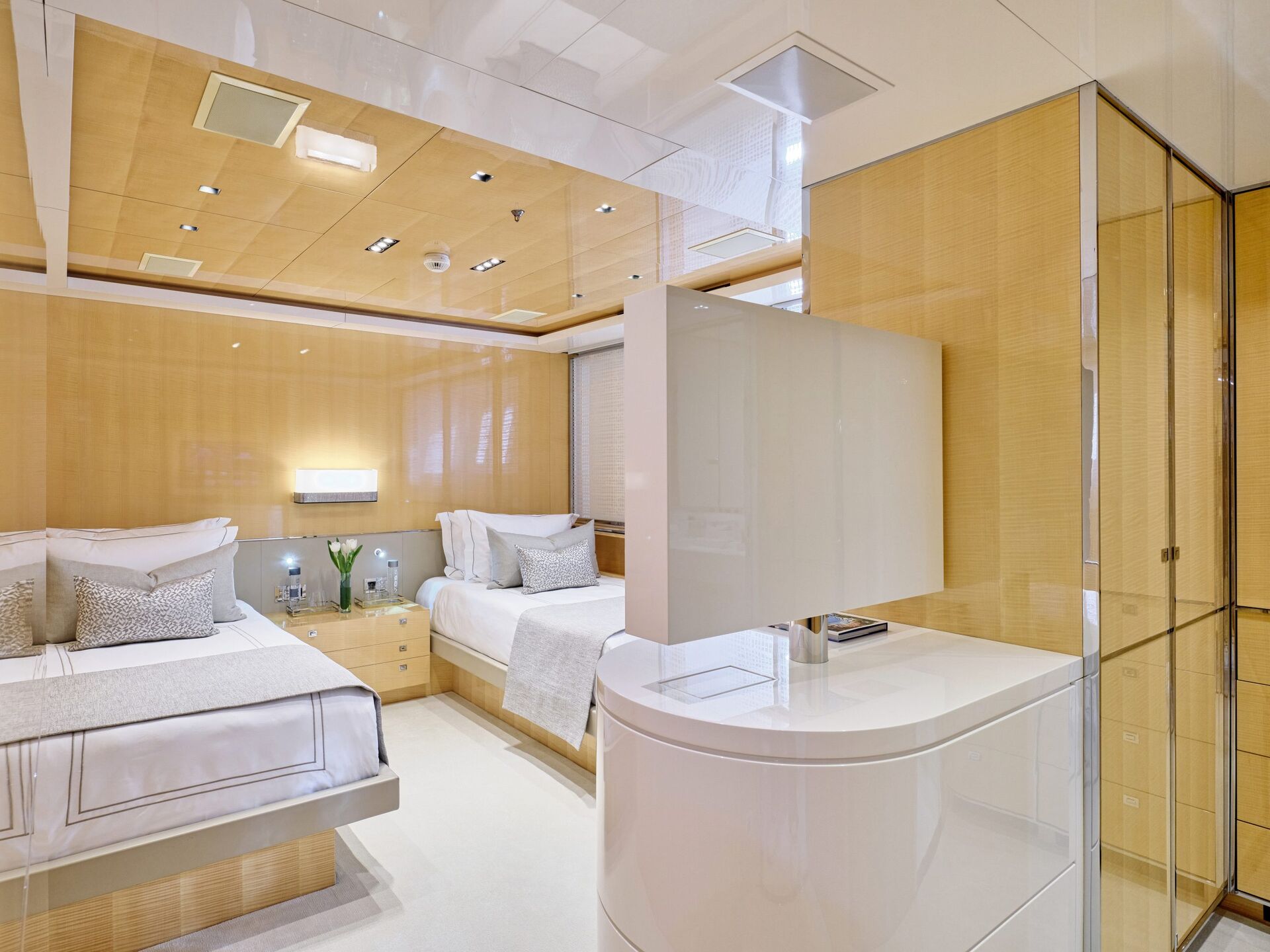 Modern bedroom with light wood paneling, two neatly made beds with white and gray bedding, built-in nightstands, mirrored wall, and a glossy white partition near the foot of the beds. Room is well-lit and minimalist.