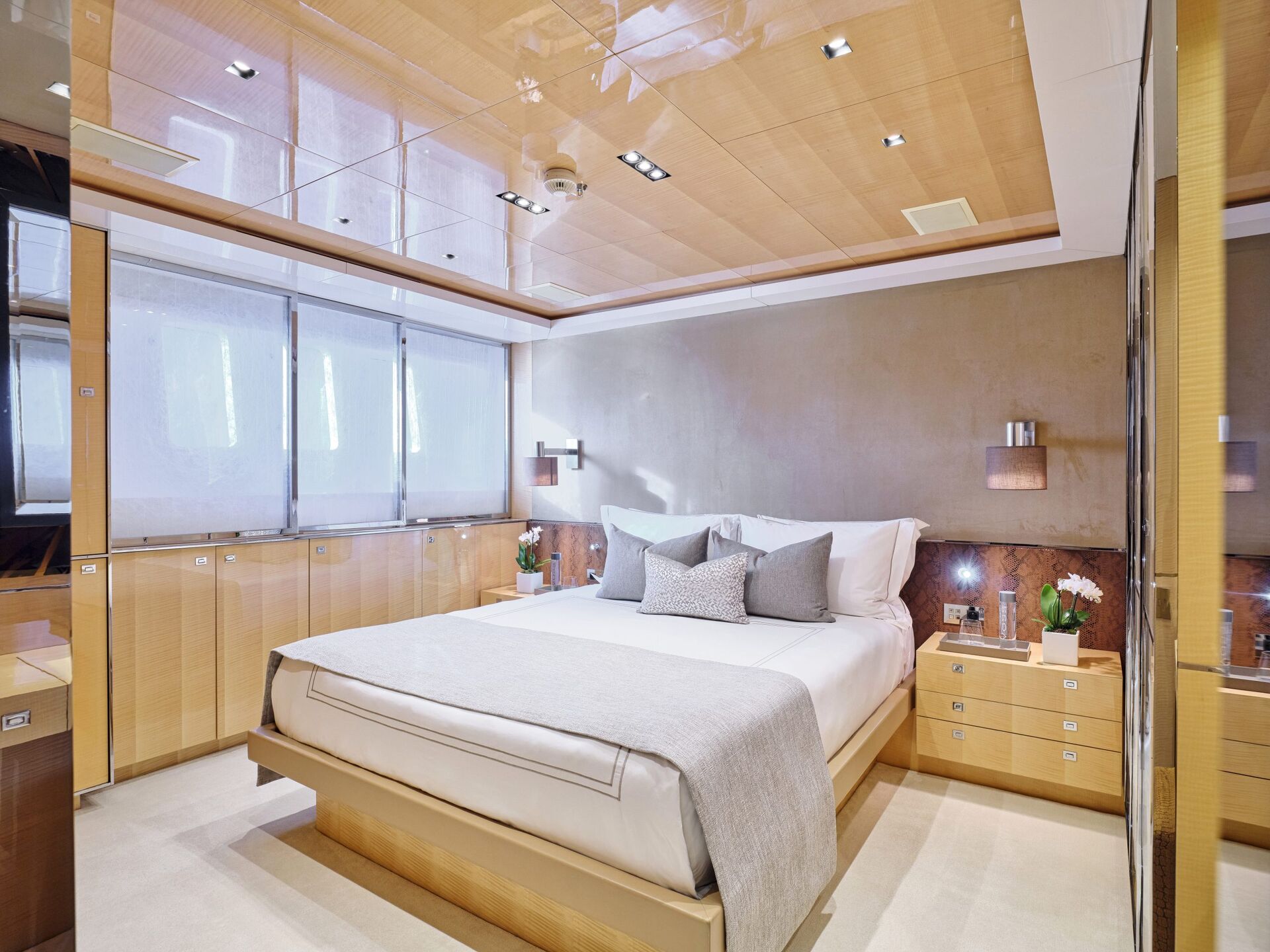Modern bedroom with light wood paneling, a large bed with white and gray bedding, built-in closets, mirrored accents, bedside tables with lamps, and potted flowers, softly lit by ceiling and wall lights.