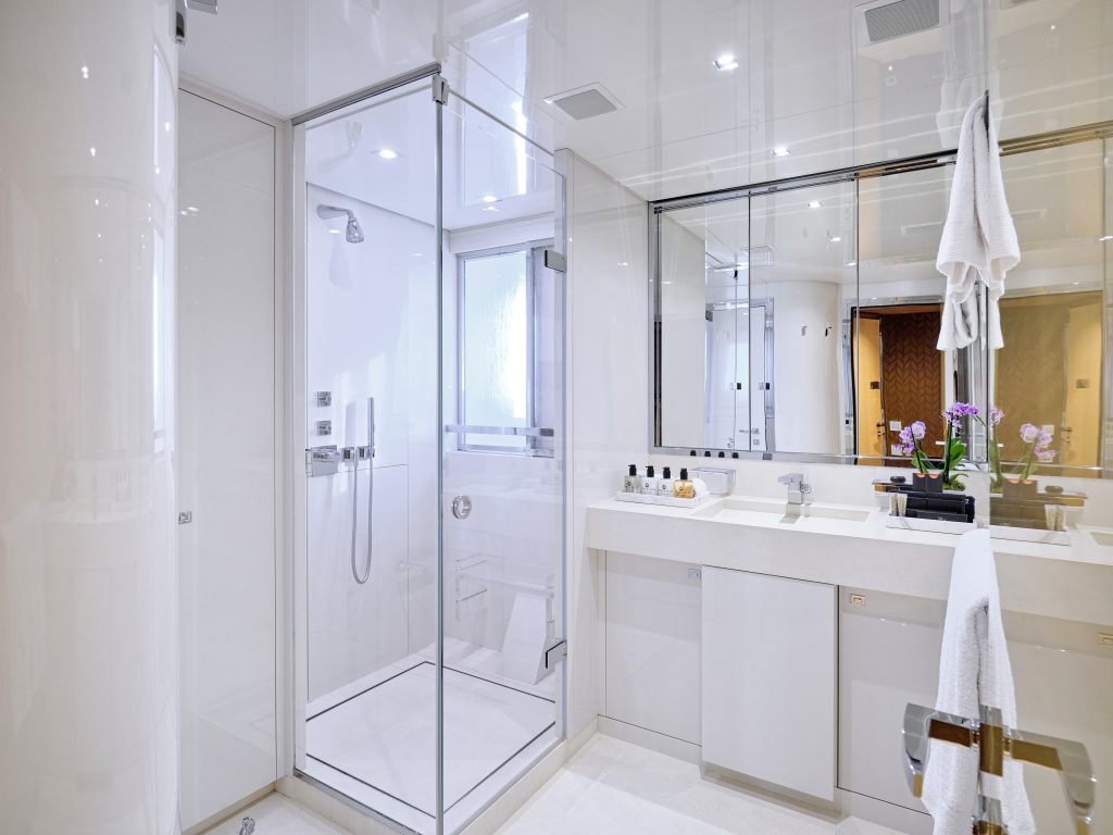 Modern bathroom with glass shower enclosure, double sinks, large mirror, white towels, and orchid plant on the counter. The room features bright lighting and a clean, minimalist design.