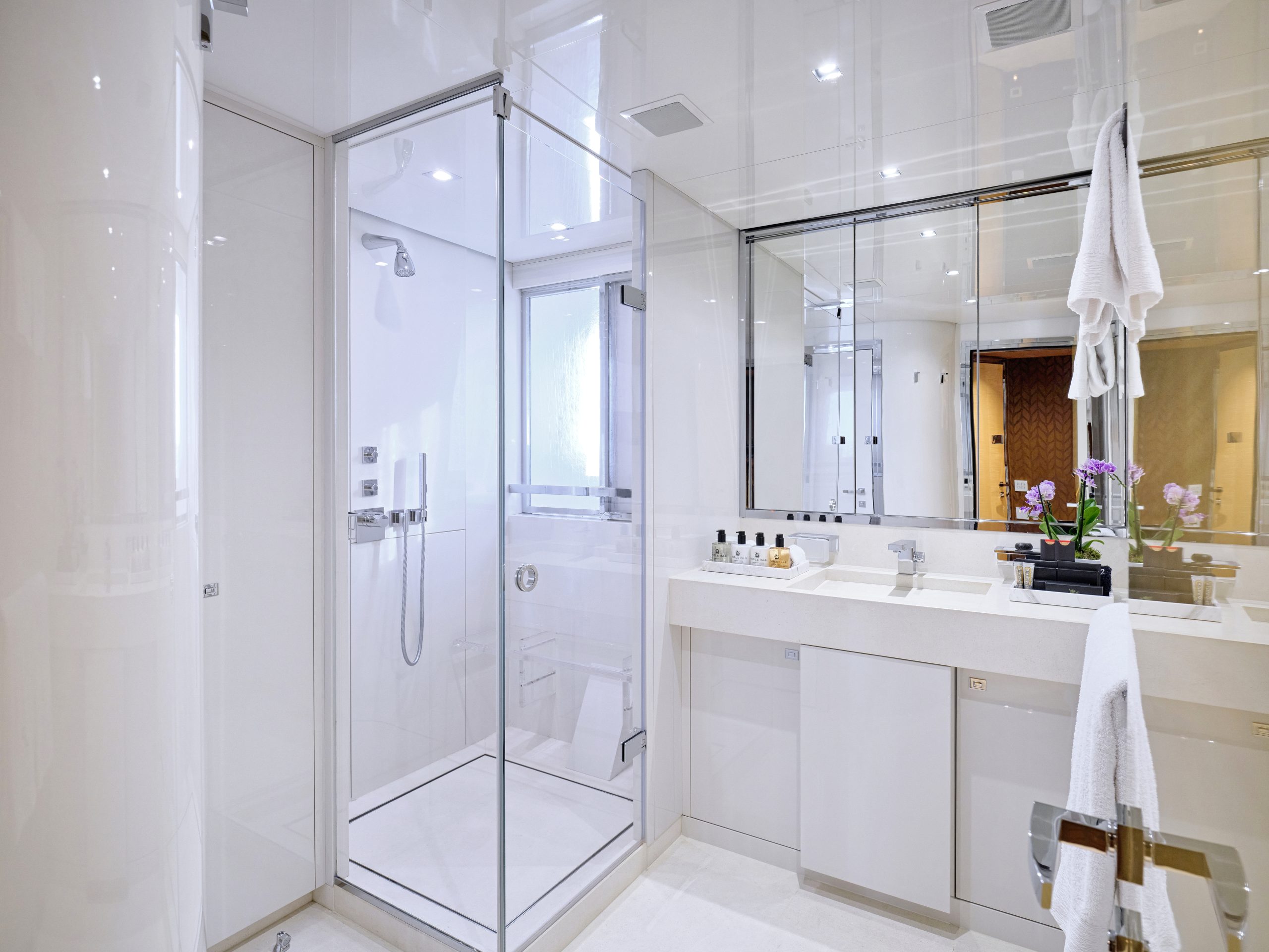 Modern bathroom with glass shower enclosure, double sinks, large mirror, white towels, and orchid plant on the counter. The room features bright lighting and a clean, minimalist design.