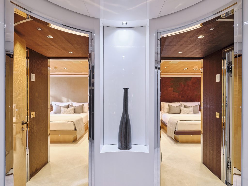 A sleek hallway with a tall black vase on a white shelf in the center, leading to two modern bedrooms with beige and brown decor, each featuring a large bed with plush pillows.