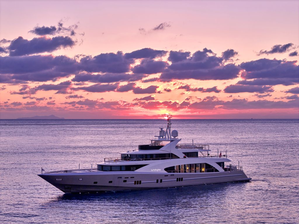 A luxury yacht sails on calm ocean waters at sunset, with the sun partially visible behind clouds and casting a pink and purple glow across the sky and sea.