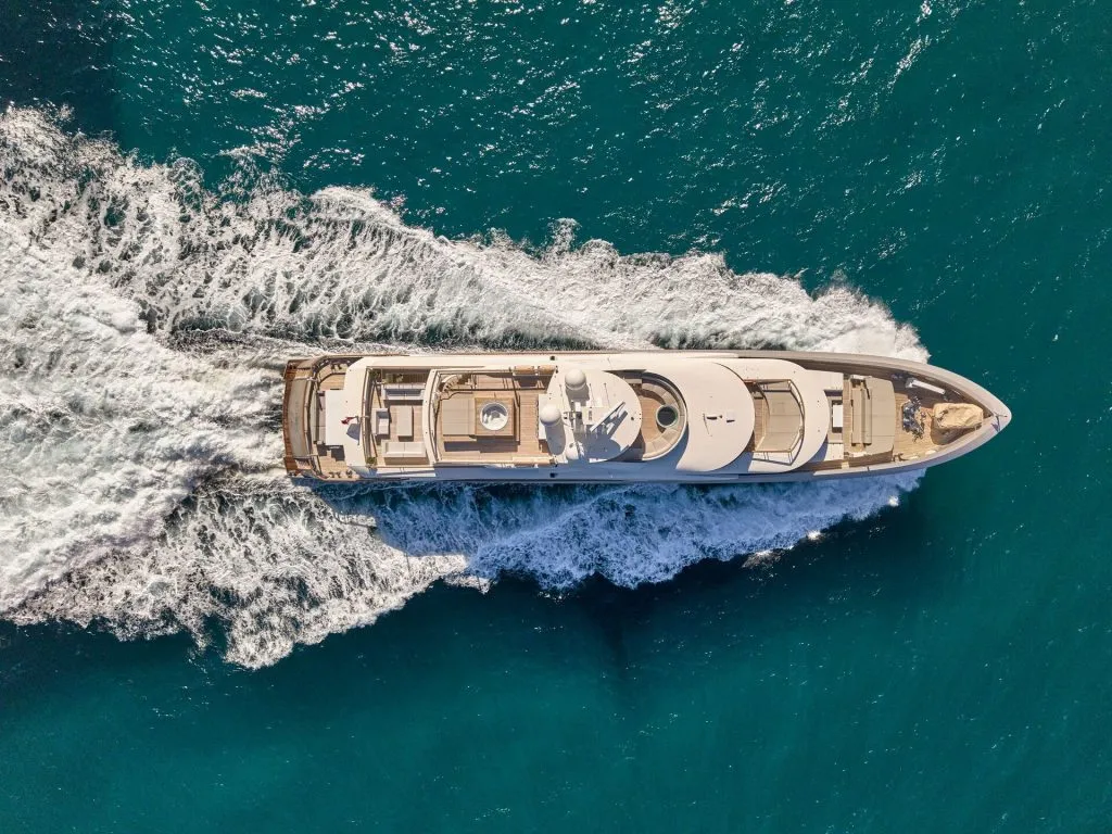 Aerial view of a luxury yacht cruising through bright blue water, leaving white waves and foam in its wake.