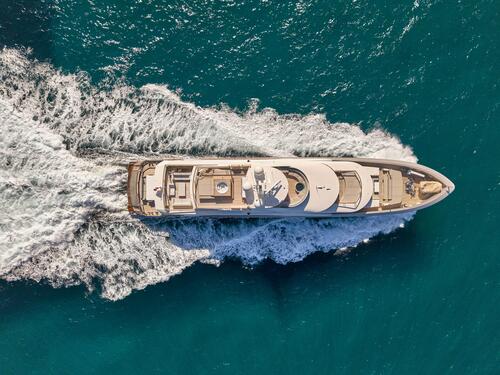 Aerial view of a luxury yacht cruising through bright blue water, leaving white waves and foam in its wake.