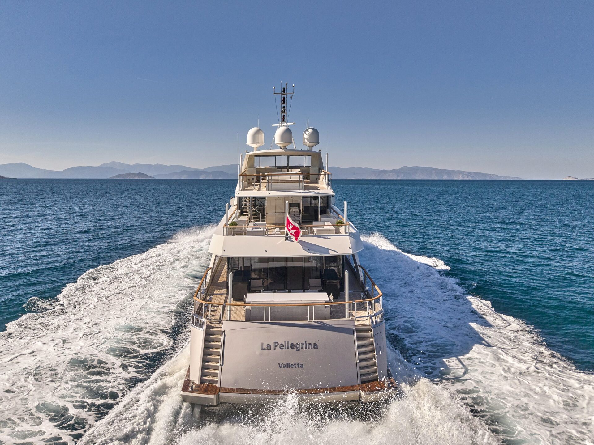A luxury yacht named La Pellegrina sails on clear blue water, leaving a white wake behind. The sky is clear with distant mountains on the horizon. A red flag flies at the yachts stern.