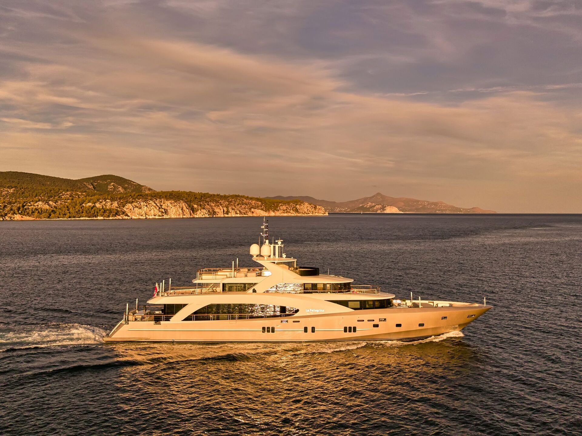 A large luxury yacht cruises on calm blue water near a rocky coastline, with hills and a partly cloudy sky in the background during sunset.