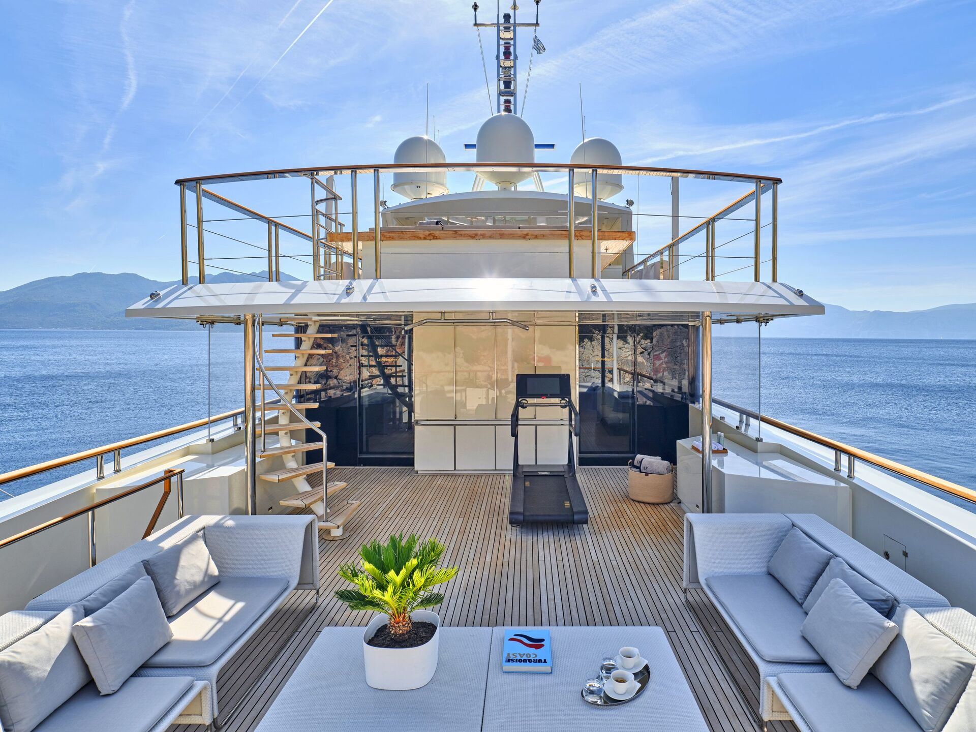 A luxurious yacht deck with modern outdoor seating, a treadmill, a small potted plant, and coffee cups, overlooking calm blue sea and distant mountains under a clear sky.