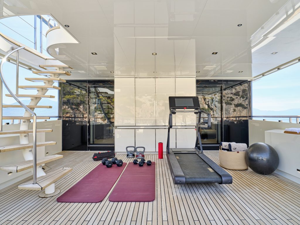 A modern yacht gym with two red exercise mats, kettlebells, dumbbells, a foam roller, a treadmill, an exercise ball, and a spiral staircase, with large windows showing a scenic ocean view.