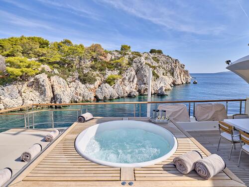 A luxury yacht deck with a round jacuzzi, rolled towels, chairs, and a table, overlooking clear blue water and rocky, tree-covered cliffs under a sunny sky.