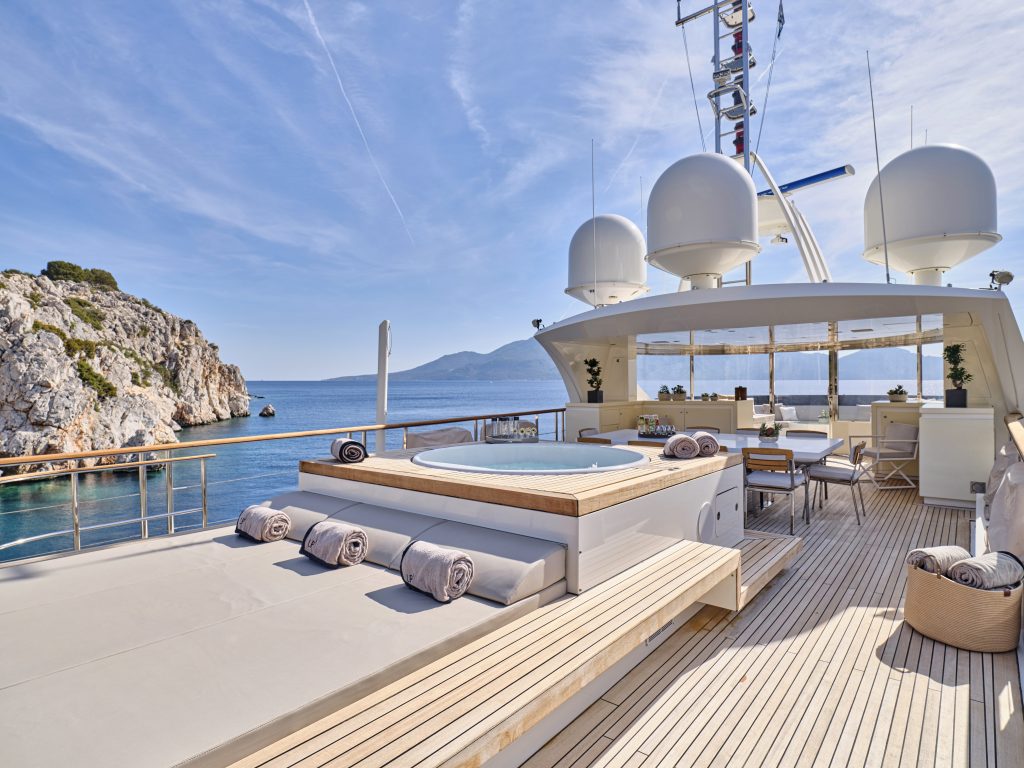 Luxury yacht deck with a hot tub, lounge chairs, and dining area, overlooking clear blue sea and rocky coastline under a sunny sky. Rolled towels are neatly placed by the tub.