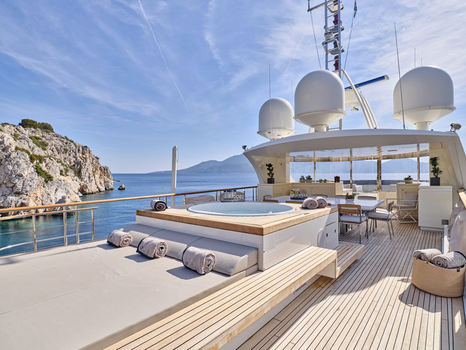 Luxury yacht deck with a hot tub, lounge chairs, and dining area, overlooking clear blue sea and rocky coastline under a sunny sky. Rolled towels are neatly placed by the tub.