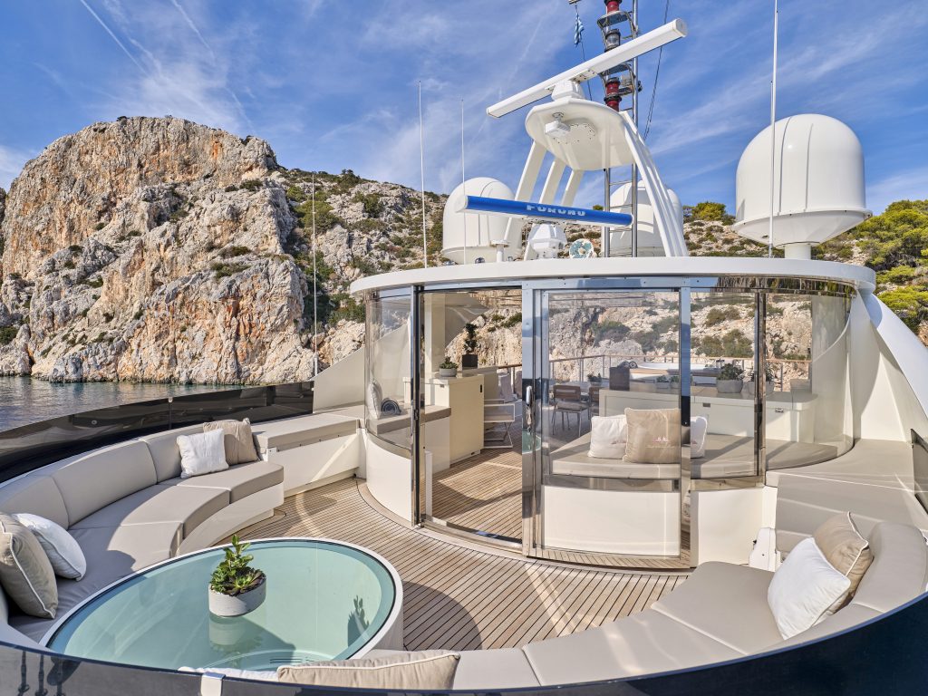 Modern yacht deck with curved seating area, glass table, and potted plant. Behind glass doors is a shaded lounge. Rocky cliffs and blue sky with wispy clouds provide a scenic backdrop on the water.