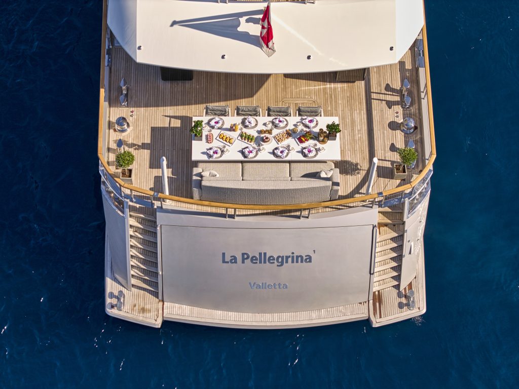 Aerial view of the stern of a luxury yacht named La Pellegrina Valletta, featuring a dining table set for a meal on a wooden deck, surrounded by blue ocean water.