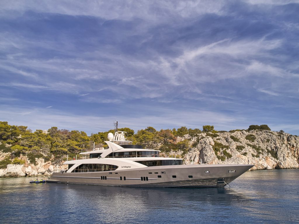 A large, modern luxury yacht is anchored near a rocky, tree-lined coastline under a blue sky with wispy clouds. The calm water reflects the yacht and surrounding landscape.
