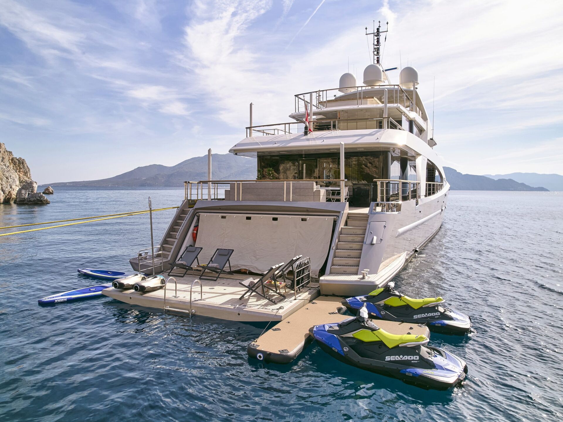 A large luxury yacht anchored in calm blue water with three Sea-Doo jet skis and paddleboards moored to a floating dock at the yacht’s rear, surrounded by scenic mountains and a partly cloudy sky.
