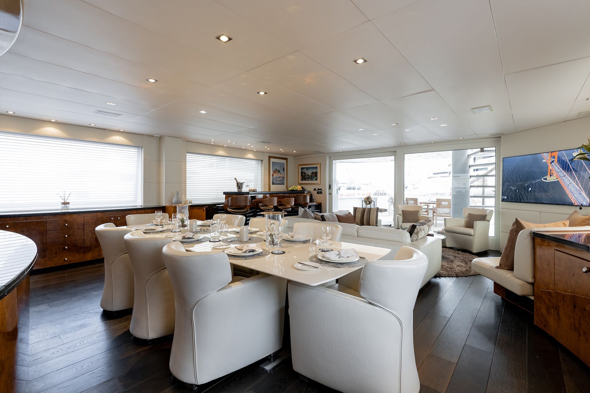 A modern yacht interior with a dining area set for six, featuring white chairs and a table, a cozy lounge with sofas, large windows, wooden flooring, and contemporary decor.