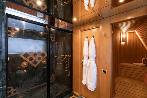 A luxurious bathroom features a glass-enclosed shower with ornate gold fixtures, two white robes hanging on wooden doors, and a wooden sauna with bench seating and rolled towels. Warm lighting enhances the cozy ambiance.