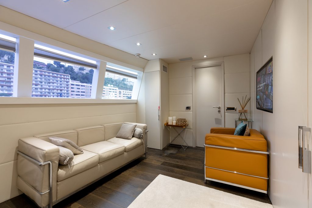 A modern yacht lounge with a beige sofa, an orange armchair, a small table with decor items, large windows, dark wood flooring, and a framed picture on the wall. Natural light fills the space.
