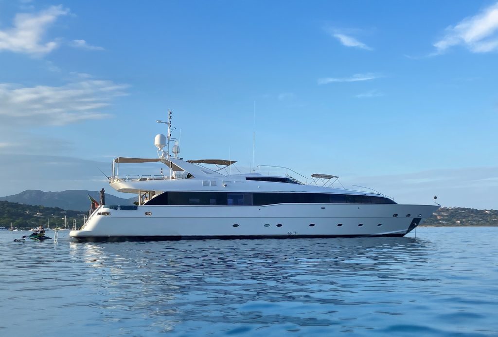 A large white motor yacht with blue accents floats on calm water under a blue sky. Hills and a distant shoreline are visible in the background.