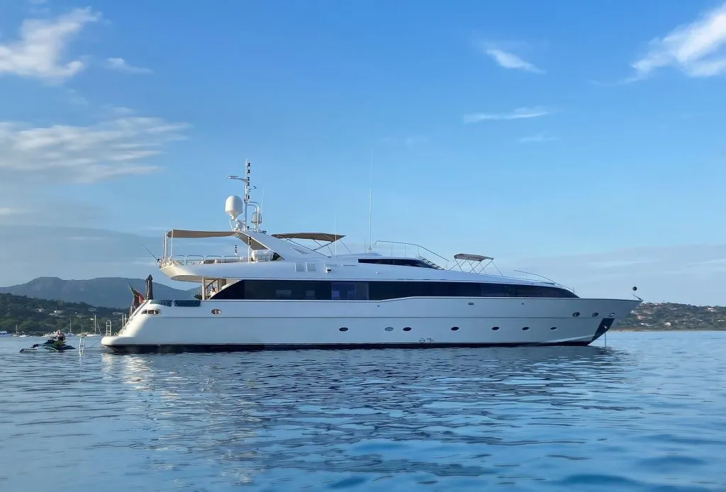 A large white motor yacht with blue accents floats on calm water under a blue sky. Hills and a distant shoreline are visible in the background.