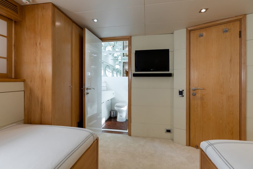 A modern, compact room with light wood paneling, featuring two single beds, a wall-mounted TV, and an open door leading to a white bathroom with a toilet and mirror.
